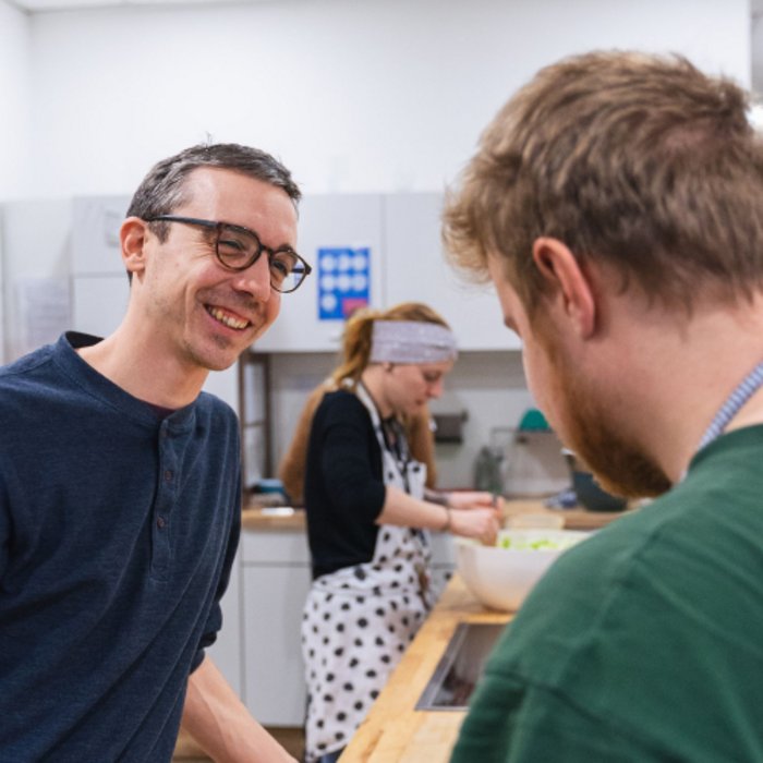Lächelnder Mann in Brille und blauen T-Shirt umgeben von zwei Personen in einer Küche.